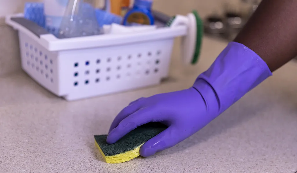 A gloved hand wiping a kitchen counter with a sponge, cleaning caddy behind